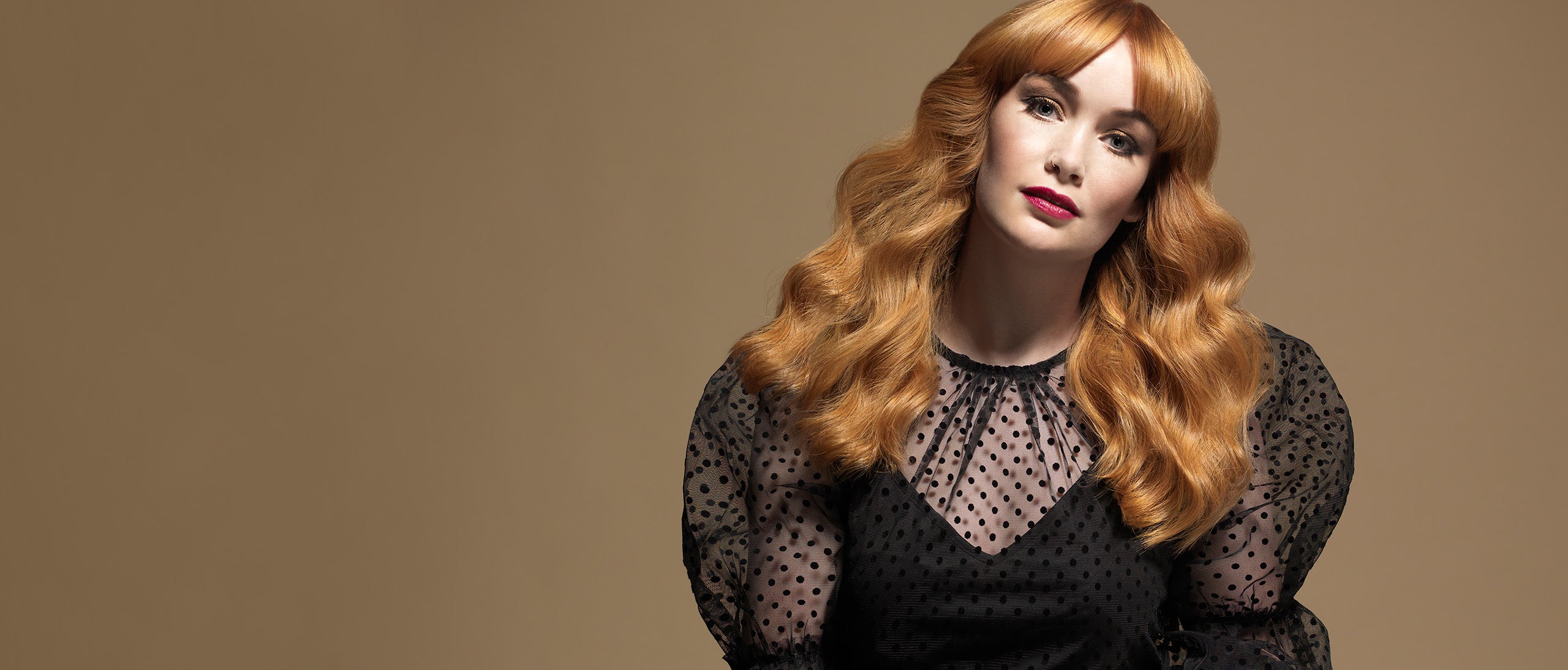 Kvinde med langt, kobberrødt bølget hår og pandehår mod beige baggrund.