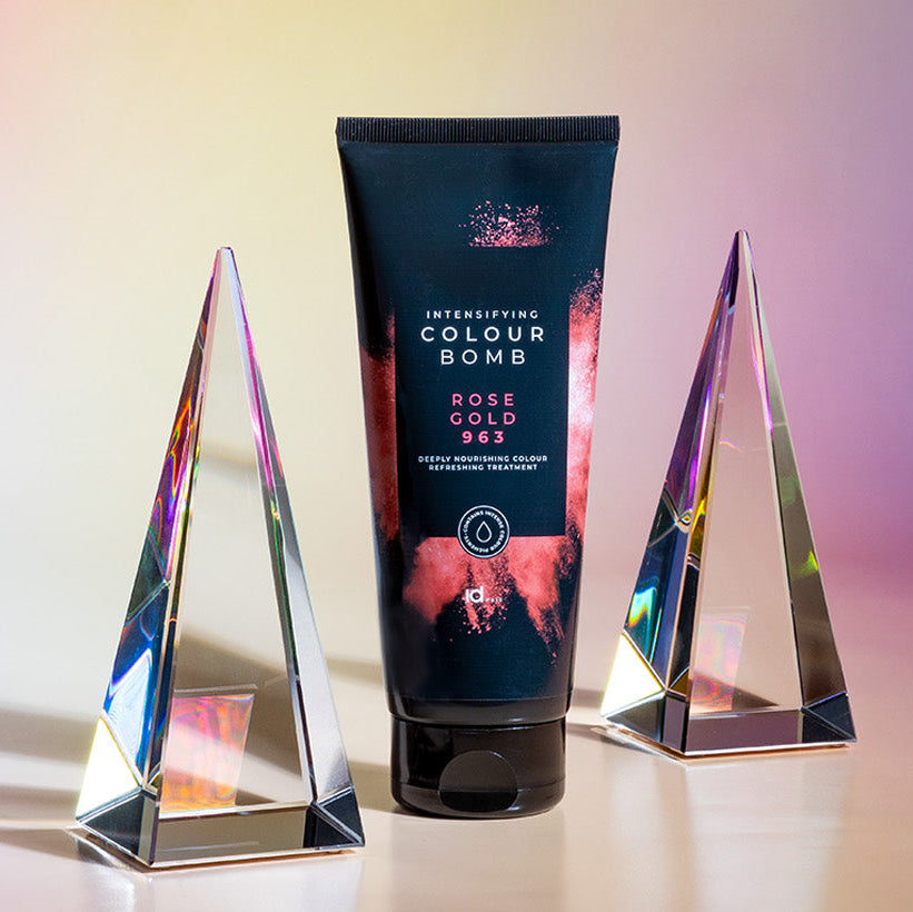 Colour Bomb Rose Gold 963 farvekur fra IdHAIR, placeret mellem to prismatiske glasdekorationer. Stilfuldt opstillet med en iriserende baggrund, der fremhæver produktets farverige og glansgivende effekt.